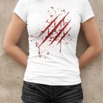 Une femme porte un t-shirt blanc avec un design graphique au centre représentant des griffures rouges ensanglantées, accompagnées de taches de sang dispersées. Le design donne un effet audacieux et accrocheur, comme si les griffures avaient déchiré le tissu. Ce t-shirt à manches courtes a une coupe ajustée, offrant un look moderne et décontracté.